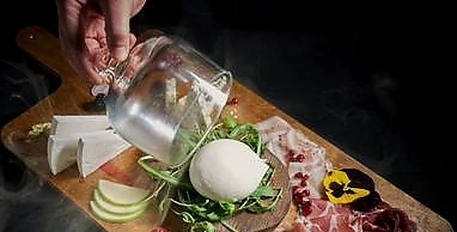 Un tagliere di salumi, burrata e formaggi viene scoperchiato da una campana di vetro affumicata.